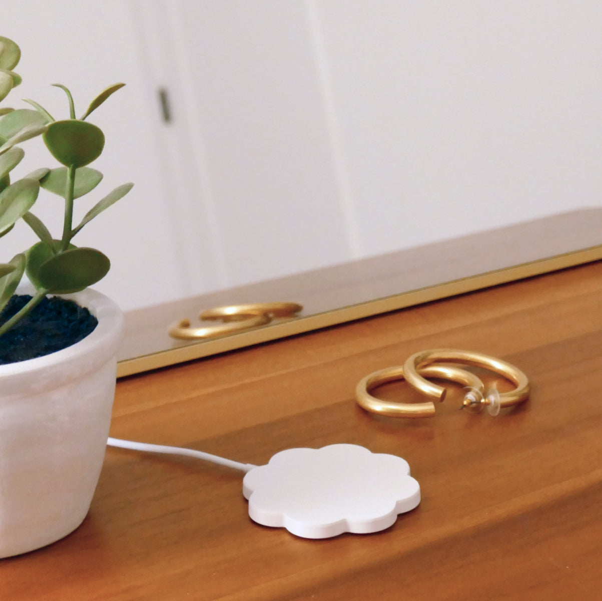 Flower shaped cover case for wireless charger on a wood, mid-century modern dresser with gold frame mirror, hoop earrings, and green plant decor